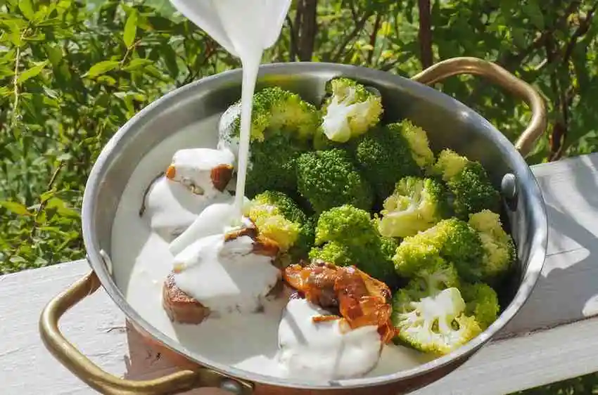 Pouring heavy cream over cooked beef and broccoli florets in a skillet to make a rich keto cheese sauce.