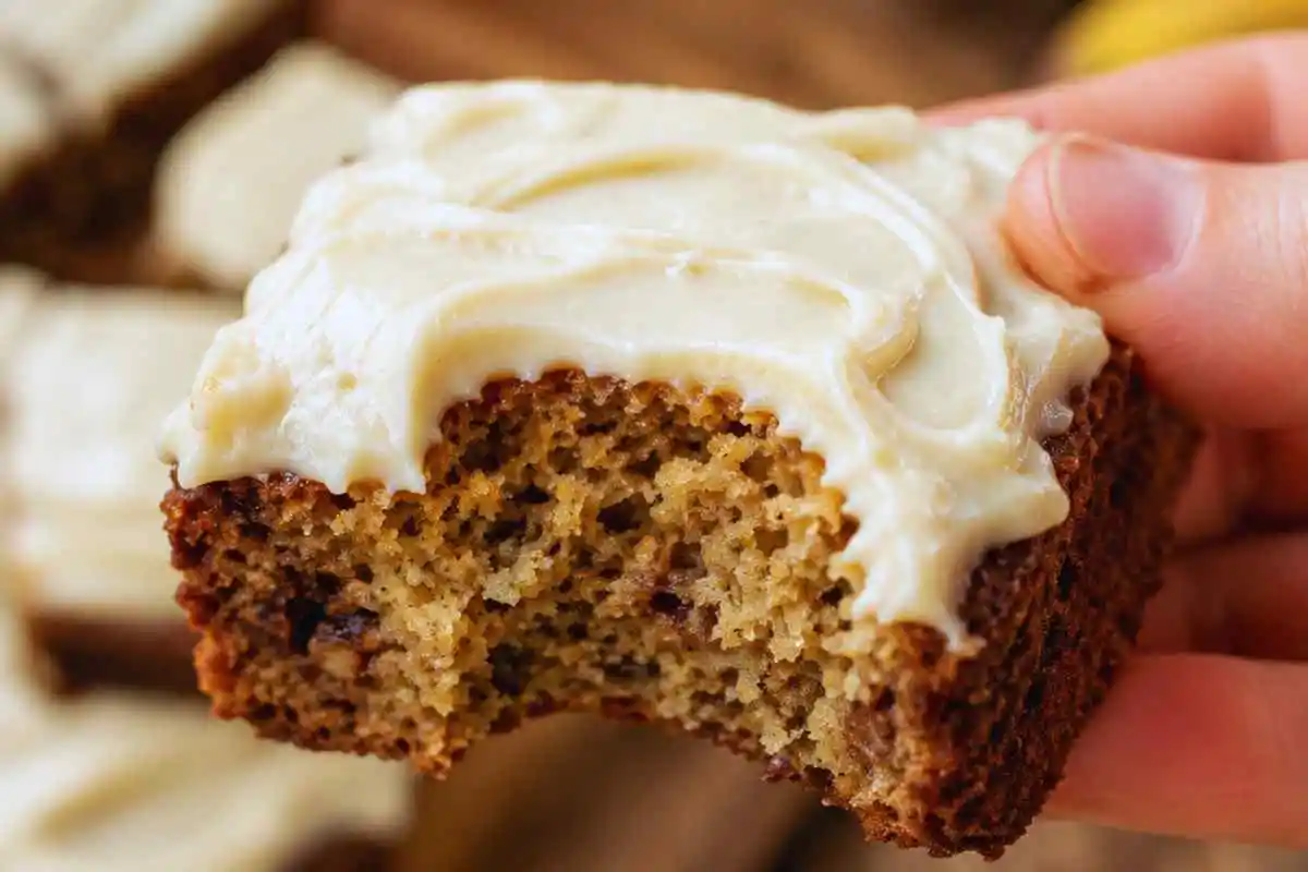 Hand holding a soft moist banana bread brownie slice with cream cheese frosting showing a bite mark.