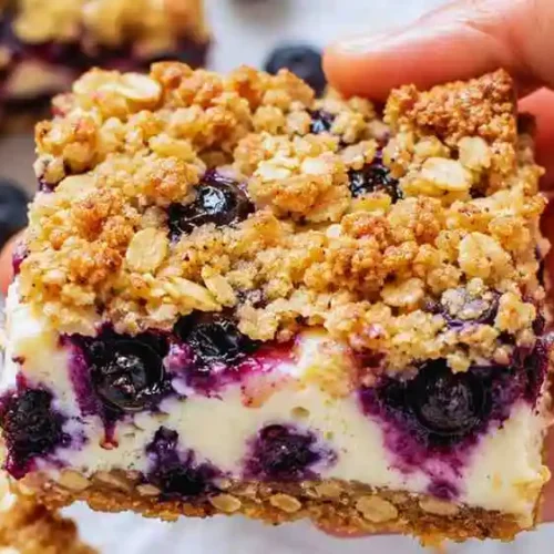 Hand holding a blueberry cream cheese bar showing the buttery oat crumble topping and rich cheesecake layer.