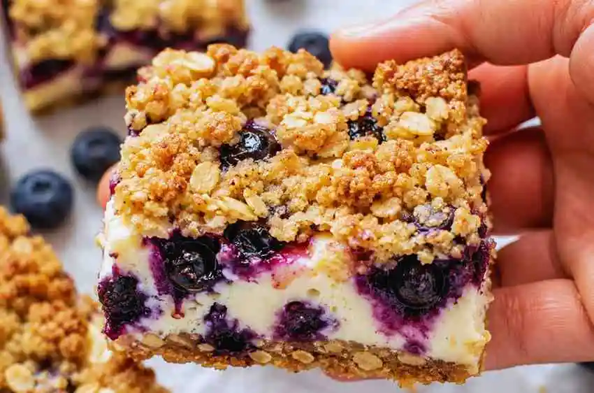 Hand holding a blueberry cream cheese bar showing the buttery oat crumble topping and rich cheesecake layer.