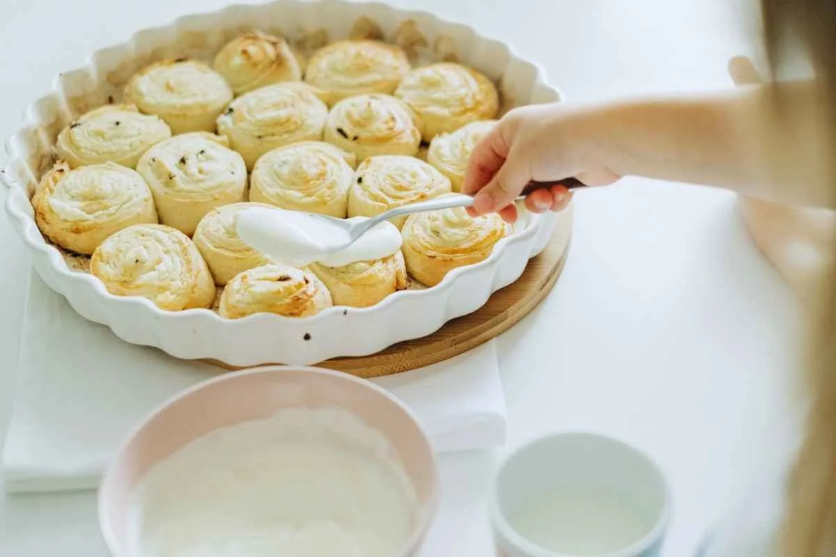 Spoon drizzling sweet white vanilla icing glaze over warm baked cinnamon rolls.