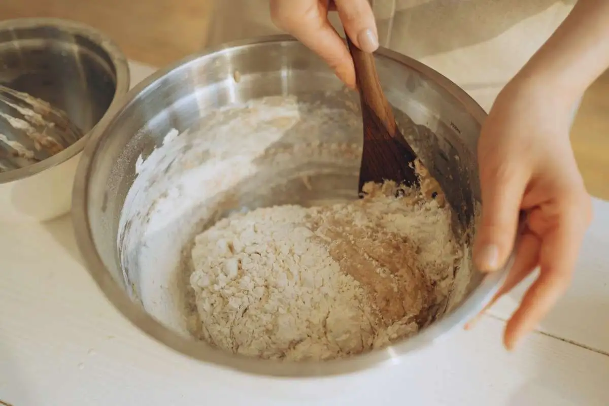 Hands mixing cake mix cookie dough in a large stainless steel bowl with a wooden spoon.