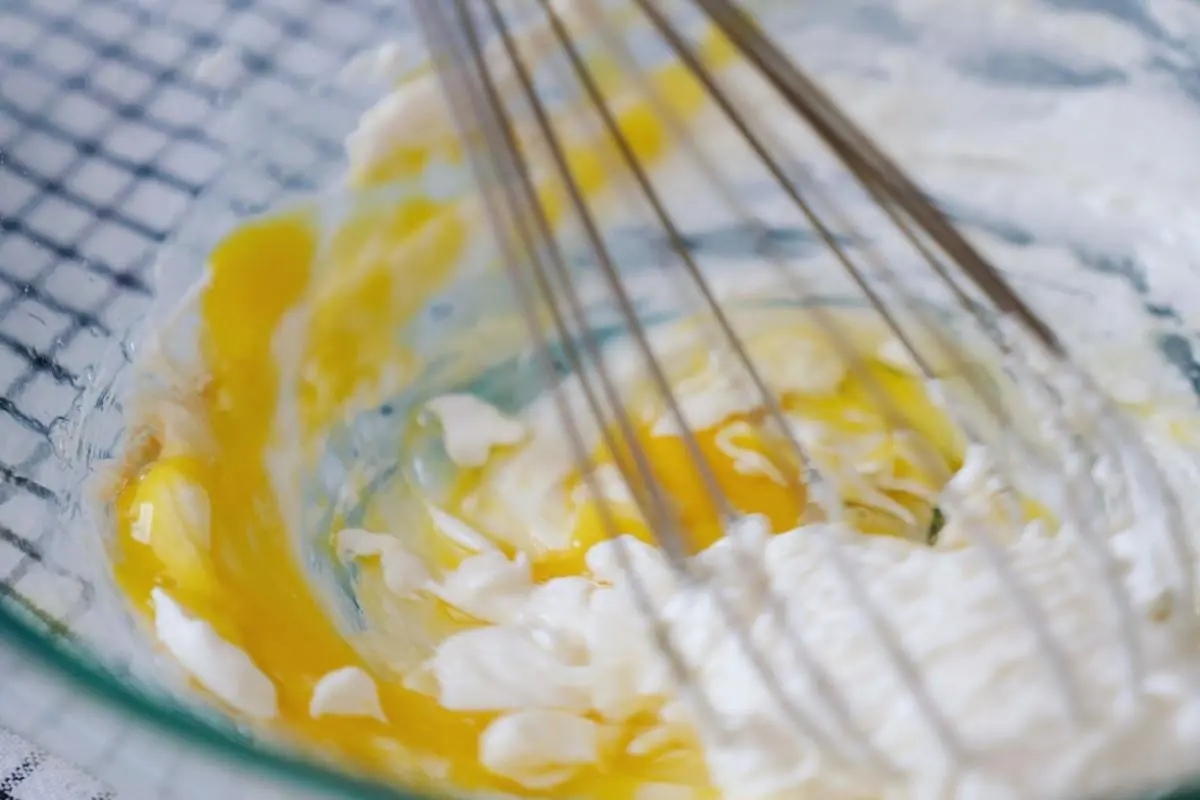 Whisking softened cream cheese and egg yolk mixture in a glass mixing bowl.