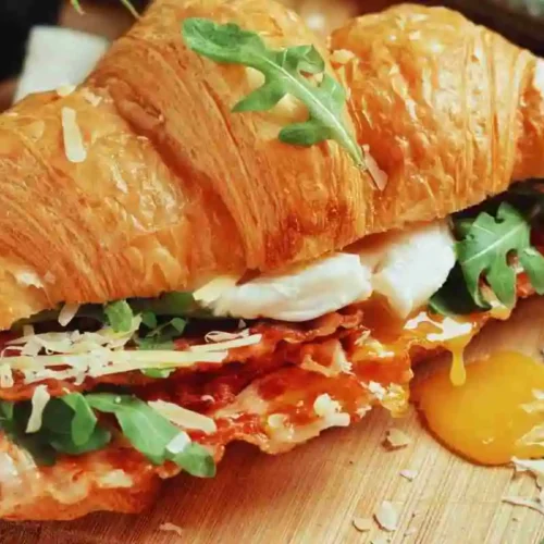 Flaky buttery breakfast croissant sandwich with crispy bacon and fresh greens on a wooden board