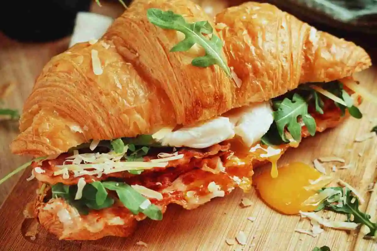 Flaky buttery breakfast croissant sandwich with crispy bacon and fresh greens on a wooden board