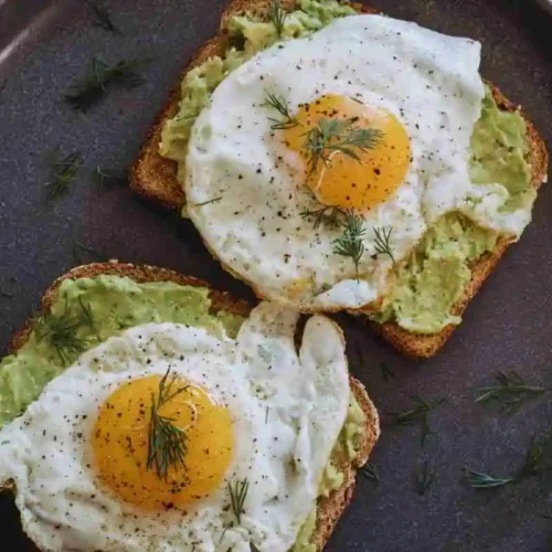Fried eggs on avocado toast served on a dark ceramic plate with fresh herbs and tomatoes