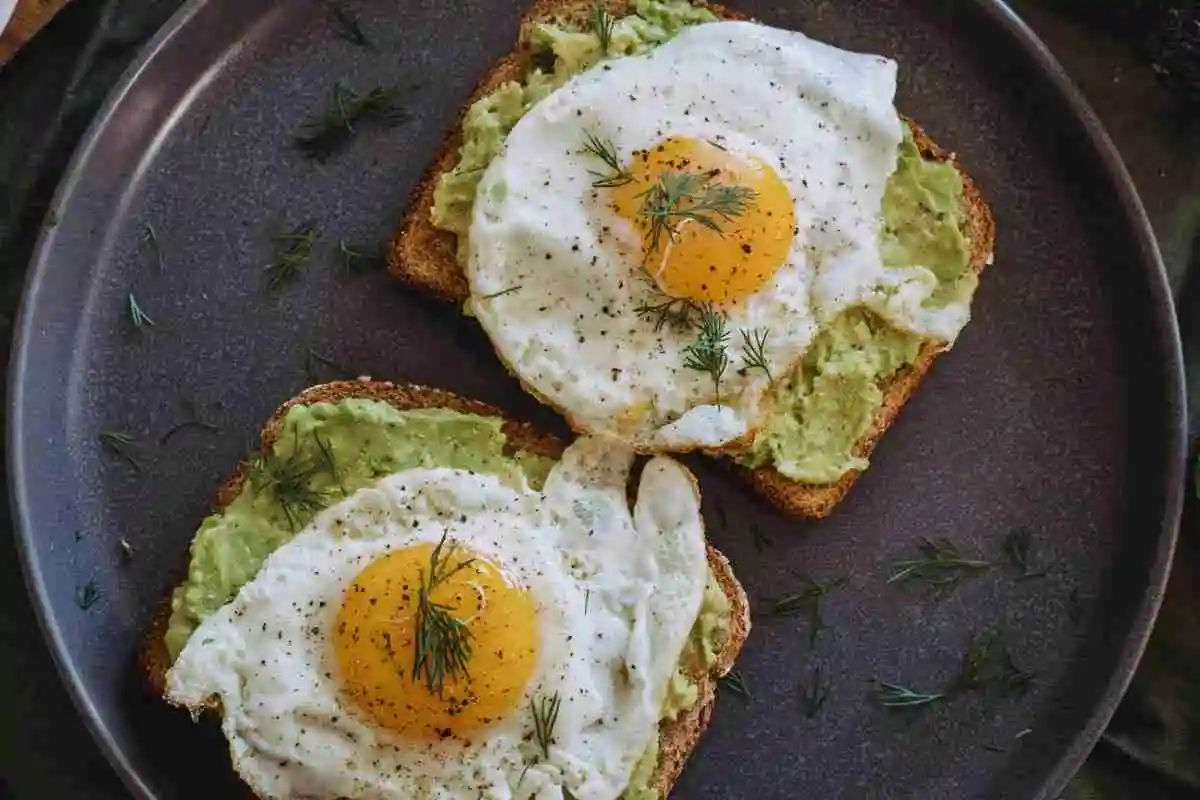 Fried eggs on avocado toast served on a dark ceramic plate with fresh herbs and tomatoes